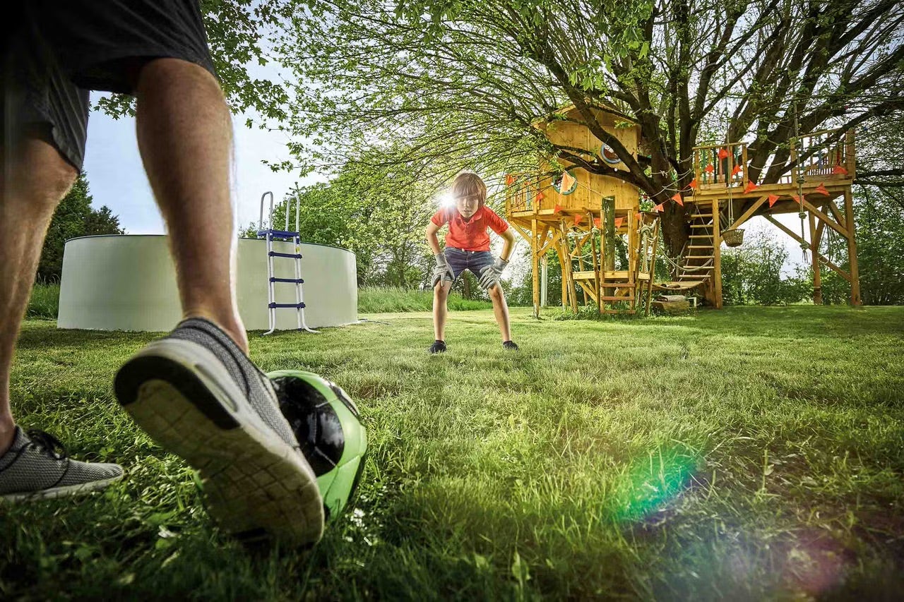 Scéna v záhrade s nadzemným bazénom, domčekom na strome a dvoma ľuďmi hrajúcimi futbal.