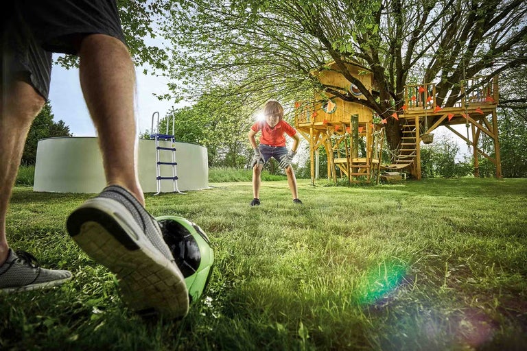 Scéna v záhrade s nadzemným bazénom, domčekom na strome a dvoma ľuďmi hrajúcimi futbal.