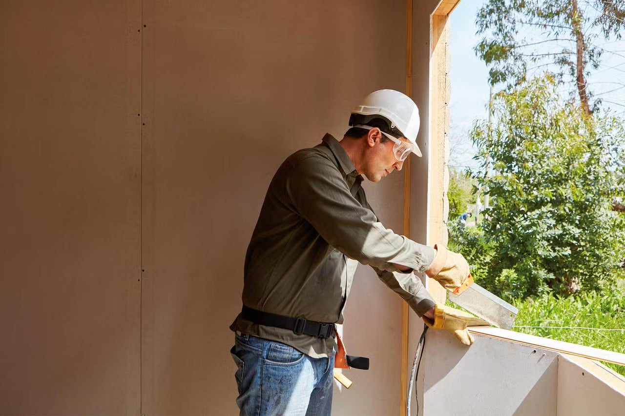 Muž s ochrannou prilbou a ochrannými okuliarmi reže drevo na okno