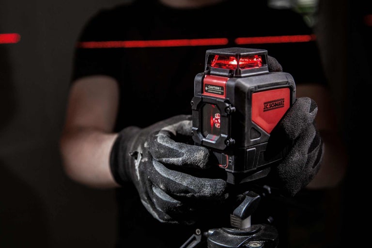 Ruka v rukavici drží laserový diaľkomer s logom JC Schwarz s červeným laserovým lúčom