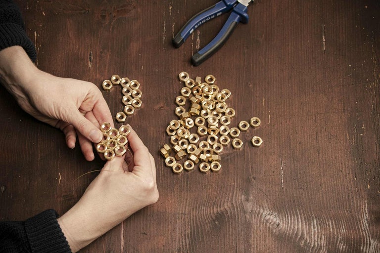 Osoba vyrába dekoráciu z matíc a drôtu, vedľa nej ležia matice a kliešte na drevenom stole.