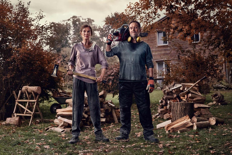 Muž s motorovou pílou cez rameno a žena so sekerou stoja pred hromadou dreva