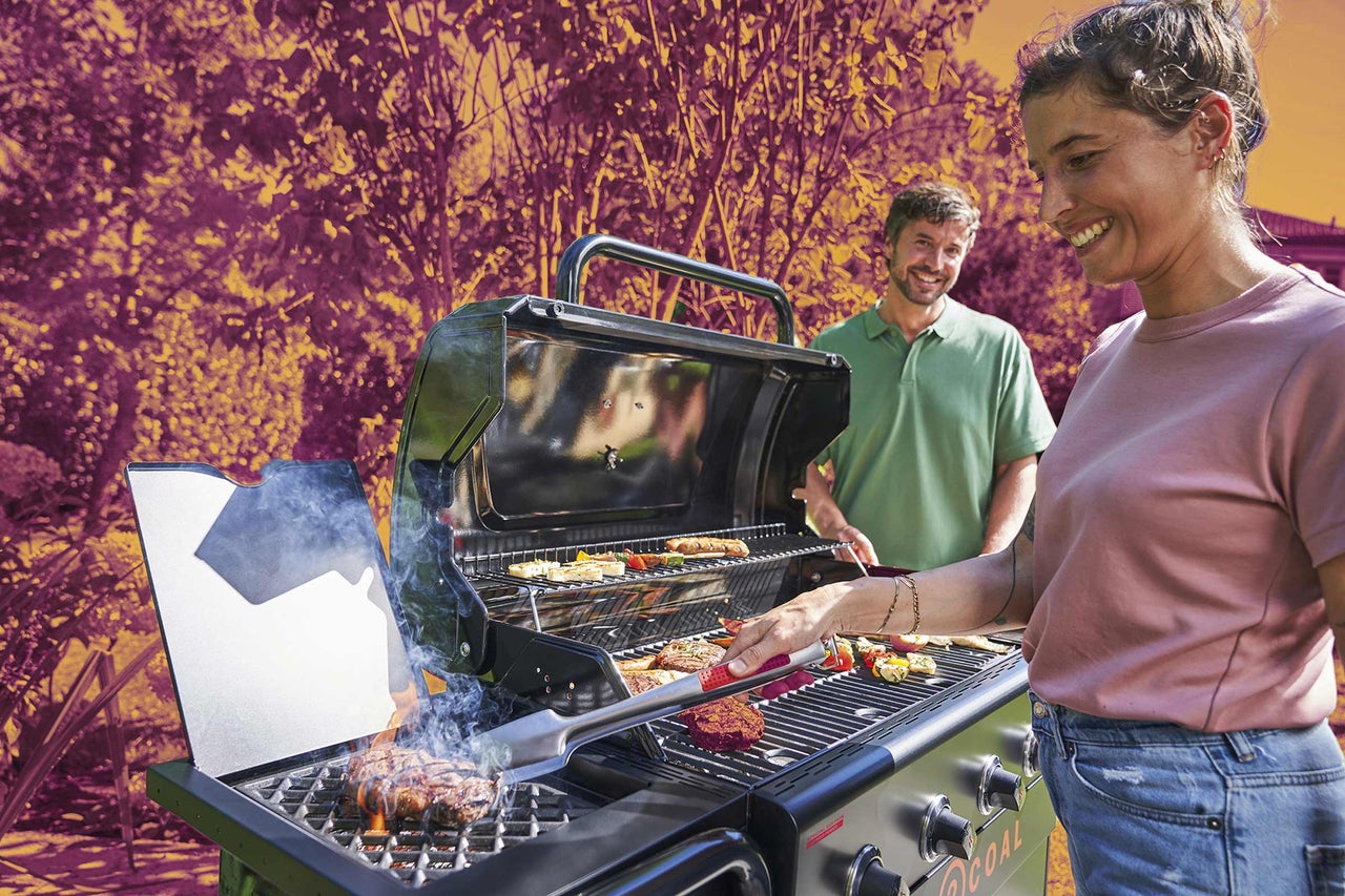 Žena griluje mäso a zeleninu na veľkom plynovom grile vo svetlej záhrade, zatiaľ čo muž v pozadí prizerá.
