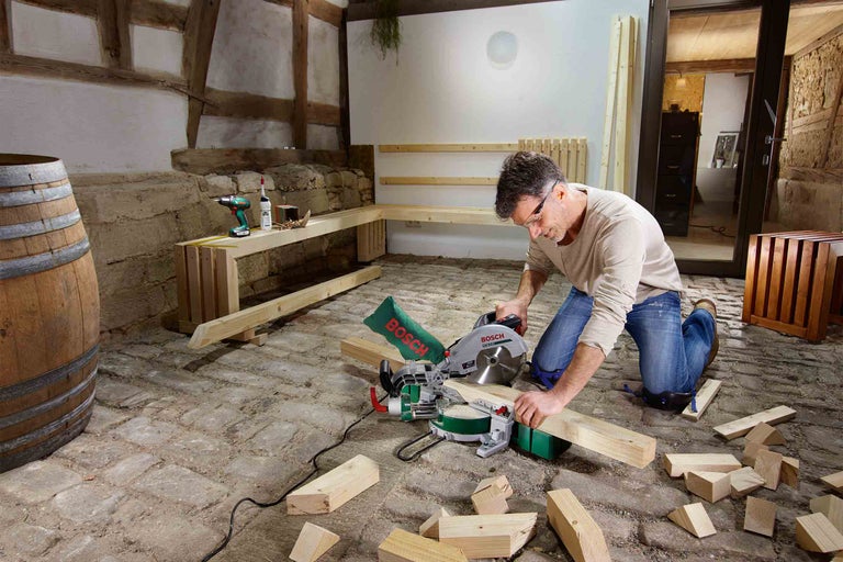 Muž reže drevo pokosovou pílou Bosch v miestnosti s drevenými trámami