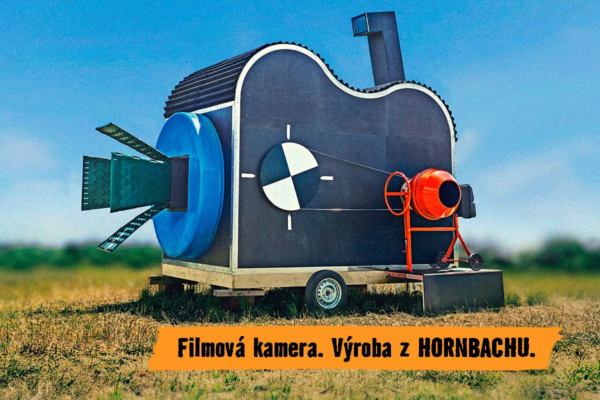 Veľká atrapa filmovej kamery na prívese vyrobená z rôznych stavebných materiálov vrátane miešačky na betón umiestnená na lúke.