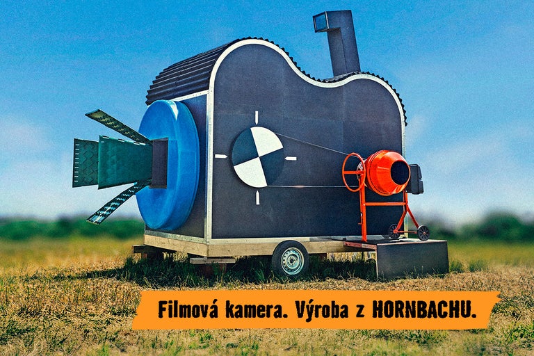 Veľká atrapa filmovej kamery na prívese vyrobená z rôznych stavebných materiálov vrátane miešačky na betón umiestnená na lúke.