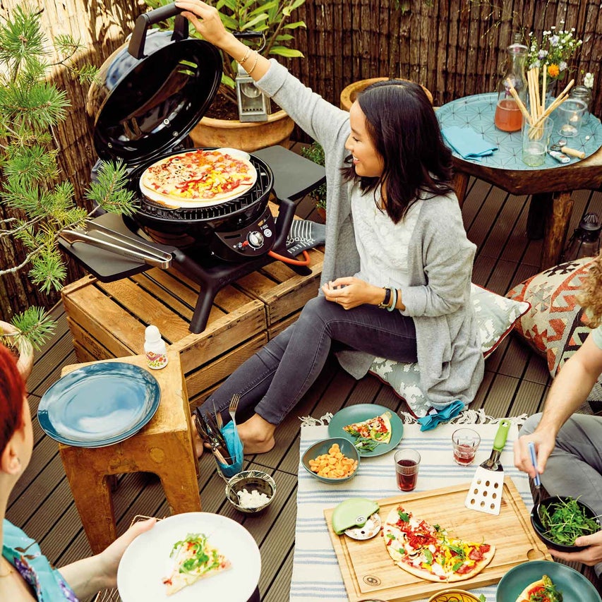 Scéna s ľuďmi, ktorí vonku jedia pizzu z grilu, s grilom, pizzou a jedálenskými potrebami na drevenej podlahe.