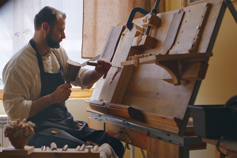 Remeselník opracováva kus dreva kladivom a dlátom na pracovnom stole.