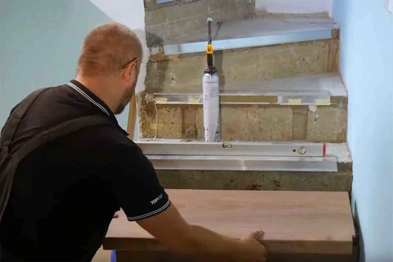 Remeselník montuje schodiskový stupeň. Je vidieť vodováha a kartuša s lepidlom.