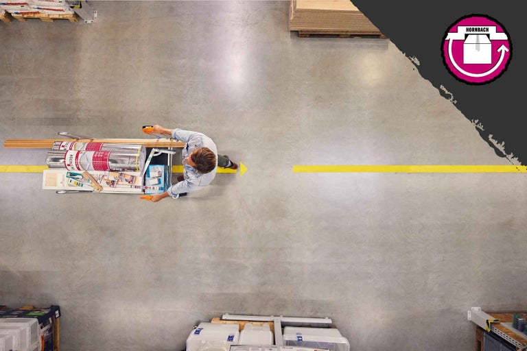Pohľad zhora na muža tlačiaceho nákupný vozík s plechovkami farieb a drevenými lištami po podlahe hobbymarketu Hornbach. Logo Hornbach