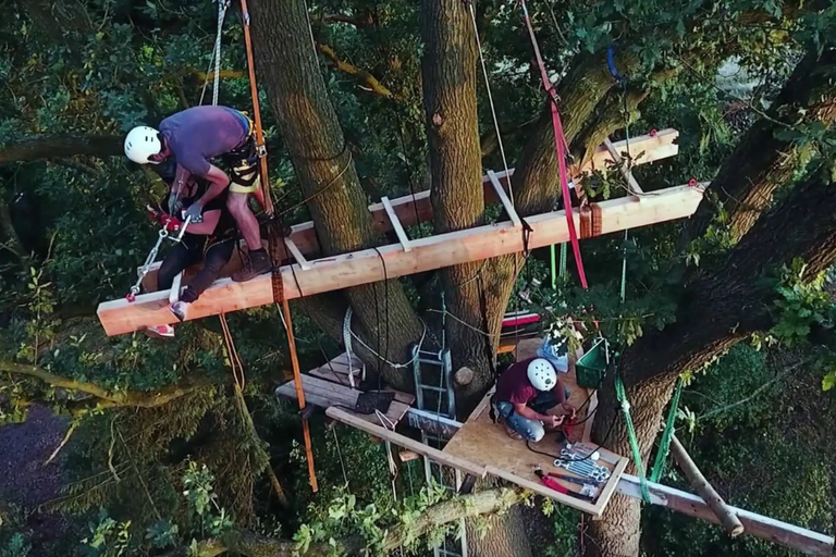 Dvaja ľudia stavajú dom na strome z drevených trámov, lán a náradia.