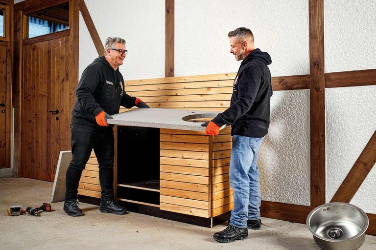 Dve osoby montujú betónovú pracovnú dosku s výrezom pre drez na drevenú spodnú skrinku.