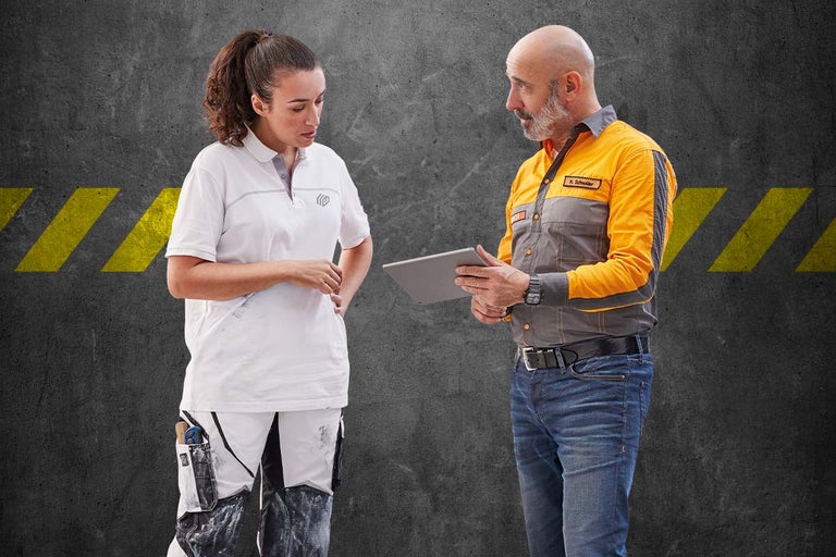 Dvaja remeselníci v pracovnom odeve si prezerajú tablet pred sivou stenou so žltými značkami.