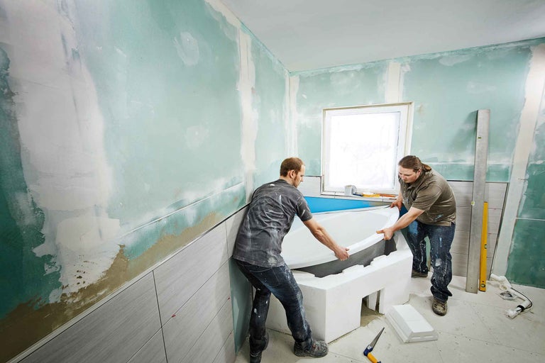 Dvaja remeselníci inštalujú vaňu v kúpeľni s nedokončenými stenami a obkladmi.