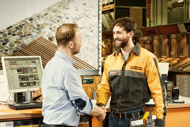 Poradca zákazníkom a zákazník si potriasajú rukami pred laminátovými výrobkami v predajni Hornbach