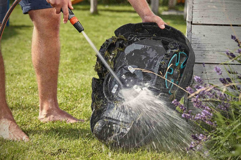 Osoba čistí robotickú kosačku záhradnou hadicou na trávniku.