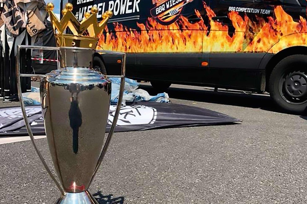 Strieborná trofej so zlatou korunou, v pozadí vozidlo s motívmi grilu