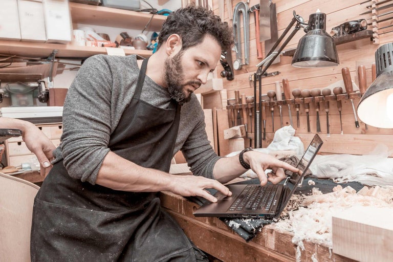 Muž so zásterou používa notebook v drevodielni, obklopený náradím a hoblinami.