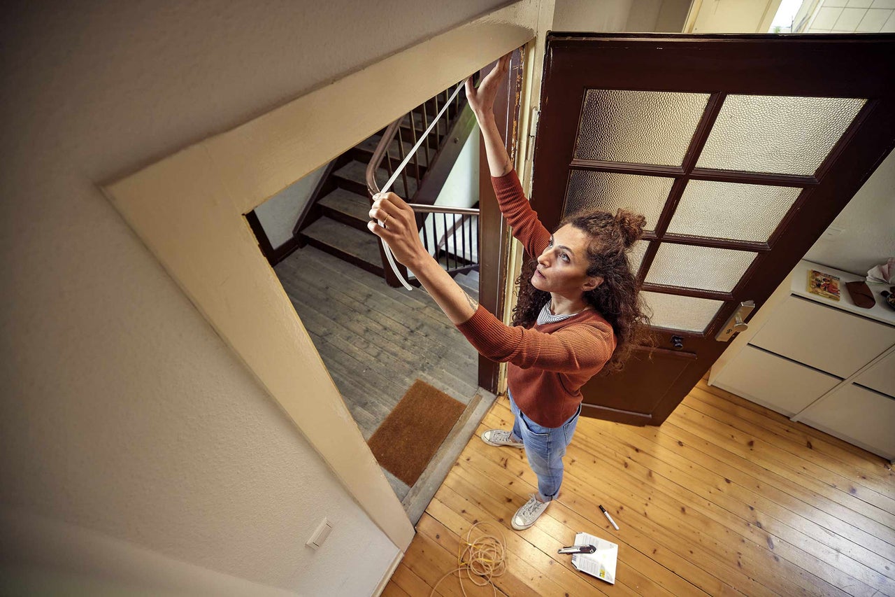 Žena meria tesniacou páskou rám dverí v dome. Na drevenej podlahe leží náradie.