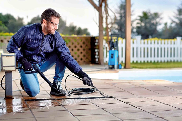 Muž čistí terasové dlaždice vonku pomocou tlakovej umývačky.