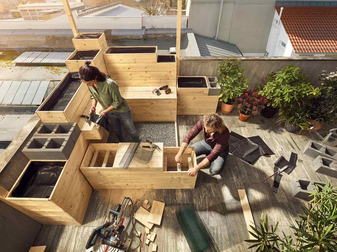 Strešná záhrada s drevenými vyvýšenými záhonmi, vybavená náradím a rastlinami, na ktorých pracujú dve osoby.
