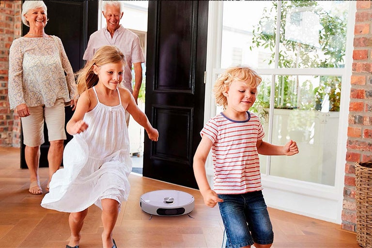 Robotický vysávač čistí drevenú podlahu, v pozadí sú vidieť deti a starí rodičia.