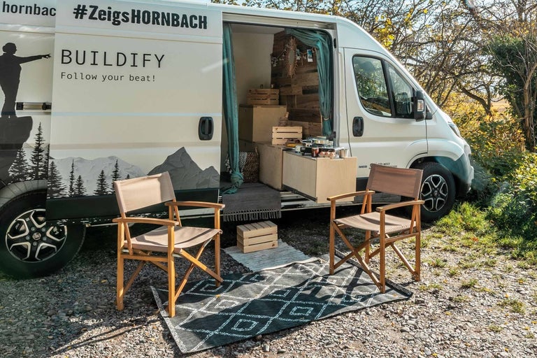 Scéna s prestavanou dodávkou, dvoma drevenými stoličkami a kobercom vonku.