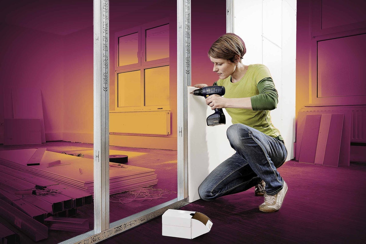 Žena pripevňuje sadrokartónovú dosku aku skrutkovačom ku kovovému rámu stojacemu na podlahe s dreveným vzorom.