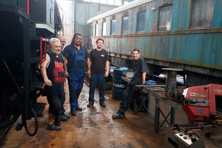 Skupina ľudí v dielni so železničnými vagónmi a náradím