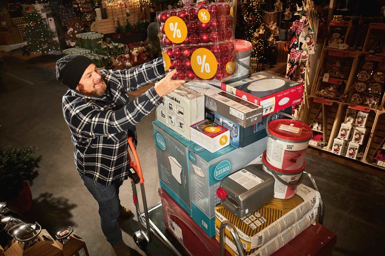 Muž v hobbymarkete Hornbach ukladá nákupy, vrátane vianočných gúľ, farby a stavebných materiálov, na vozík.