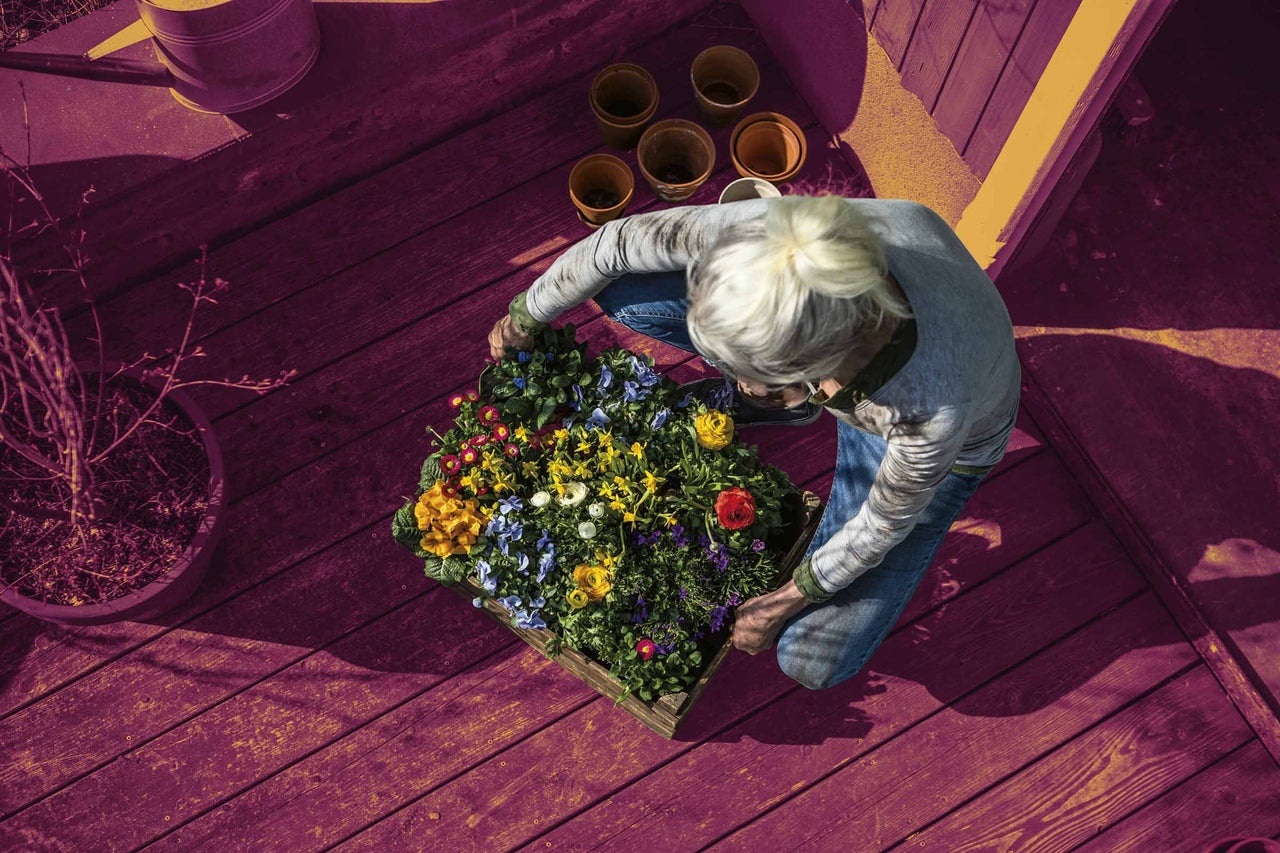 Žena kľačí na drevenej terase a aranžuje kvety do drevenej debničky, okolo ktorej sú kvetináče a krhla.