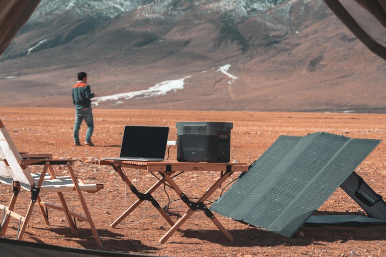 Kempingová scéna s prenosnou elektrárňou, solárnym panelom a notebookom na kempingovom stole s horskou scenériou v pozadí