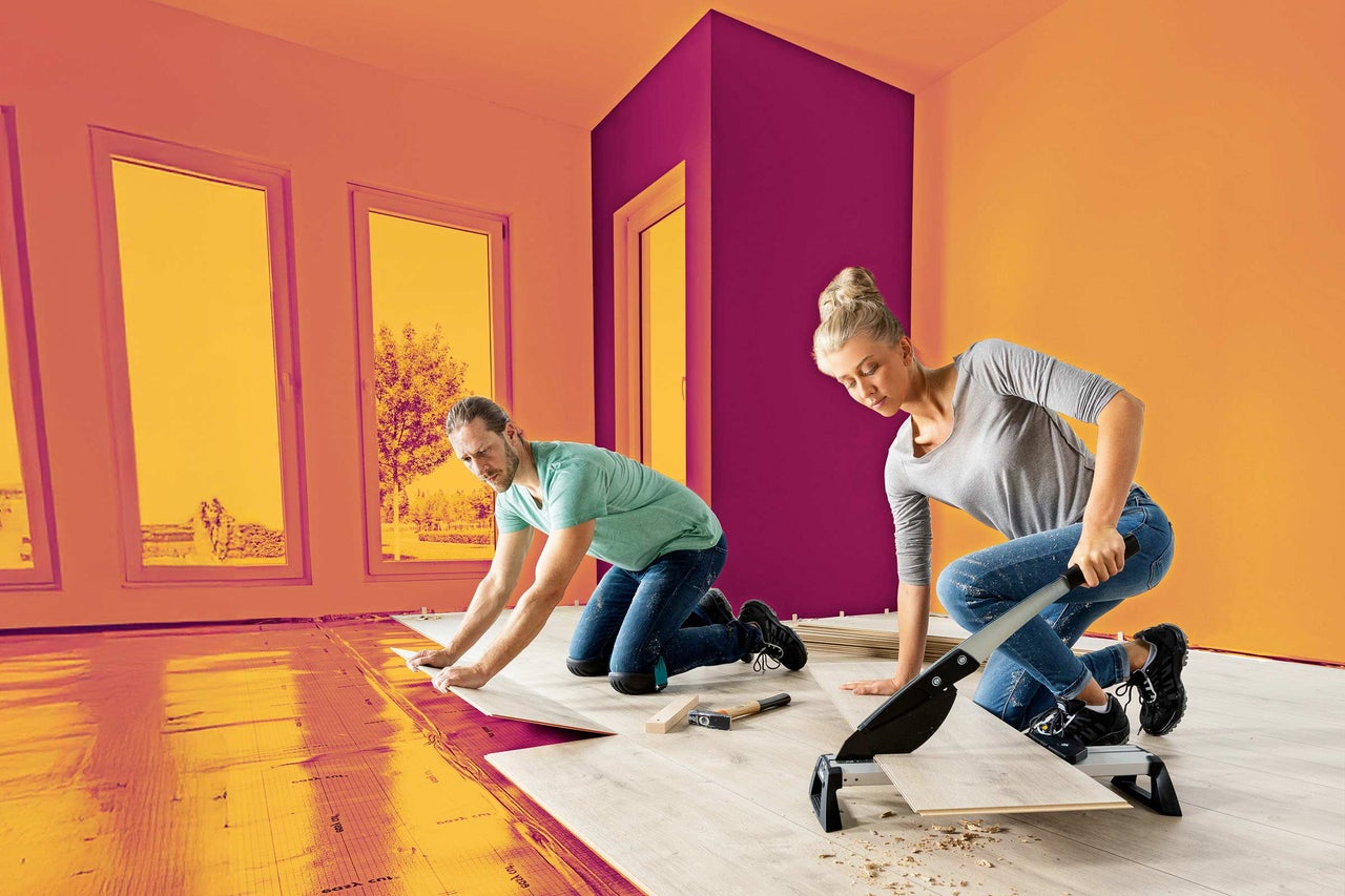 Dve osoby kladú laminát v miestnosti s podložkou a rezačkou laminátu.
