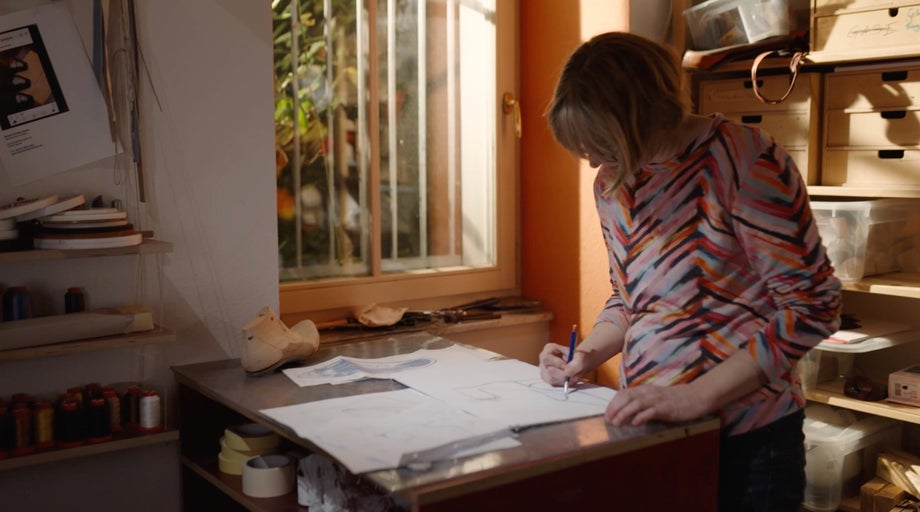 Žena navrhuje topánky na stole v svetlej miestnosti s policami a oknom.