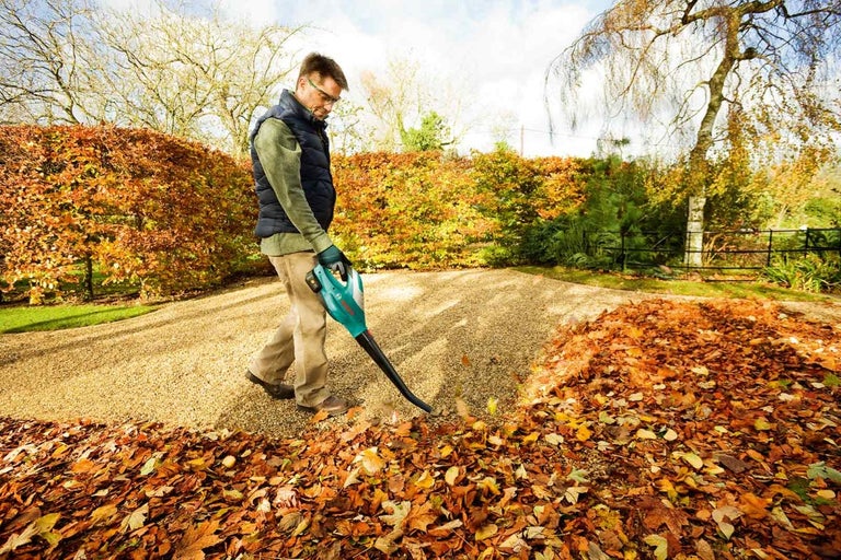 Muž používa fúkač lístia v záhrade plnej jesenného lístia.