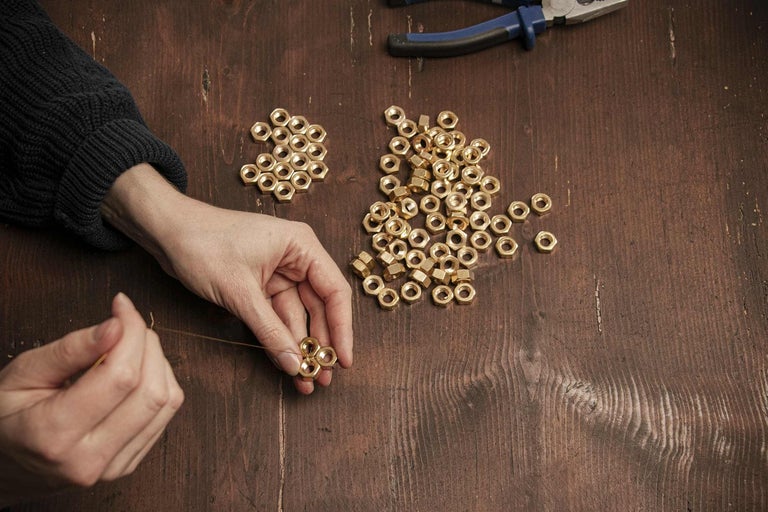 Detailný záber osoby spájajúcej mosadzné matice drôtom na drevenom stole. V pozadí ležia kľúče.