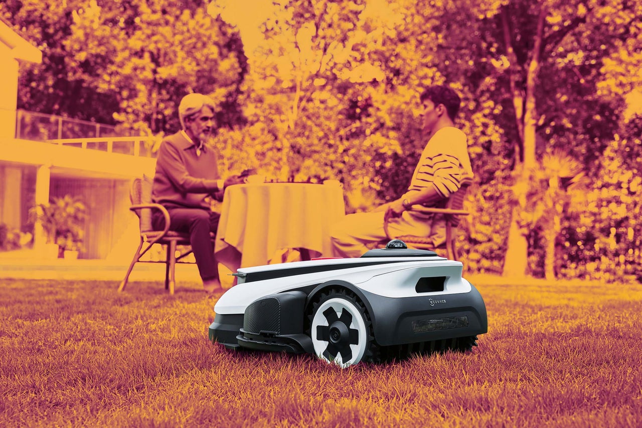 Robotická kosačka Ecovacs na trávniku v záhrade, v pozadí sedia dve osoby pri stole.