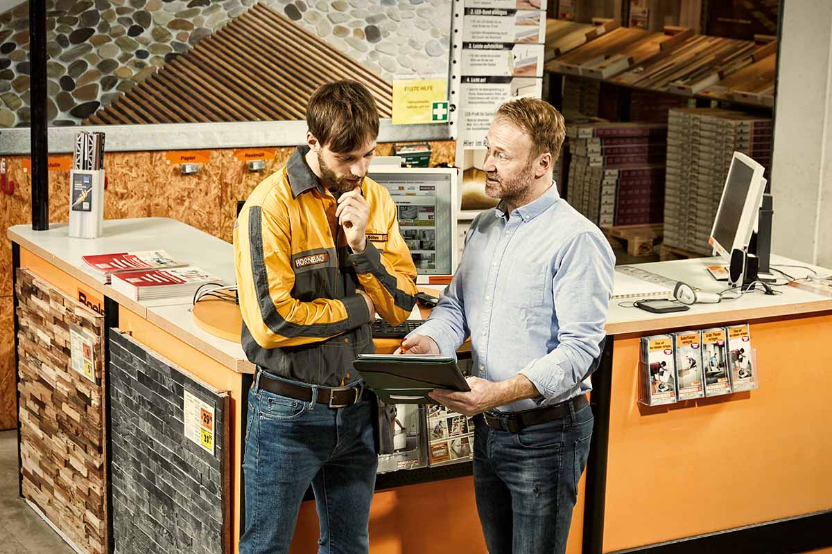 Zamestnanec Hornbachu v uniforme radí zákazníkovi pri servisnom pulte v hobbymarkete pri prezeraní tabletu.