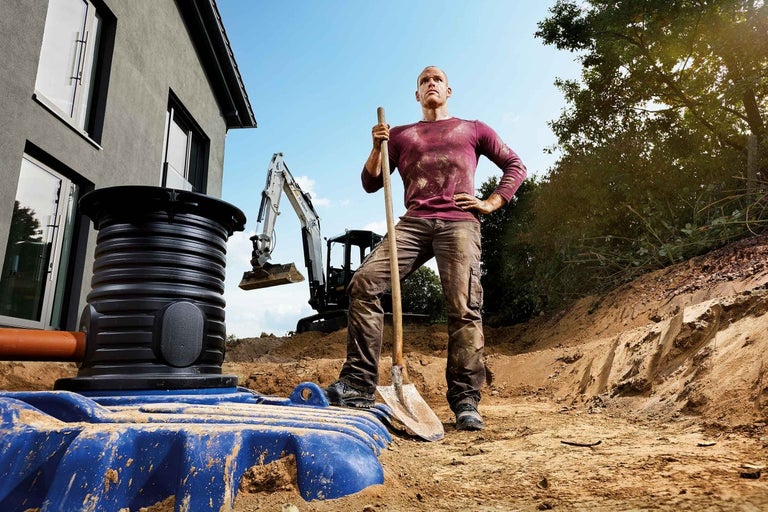 Scéna s mužom s rýľom, bagrom a zemnou nádržou na stavenisku pred domom