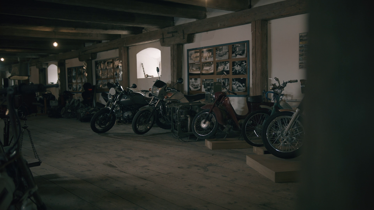 Rad motocyklov v hale s drevenou podlahou a stropom s drevenými trámami.