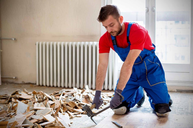 Muž v pracovnom odeve odstraňuje drevenú podlahu pomocou kladiva a dláta v miestnosti.