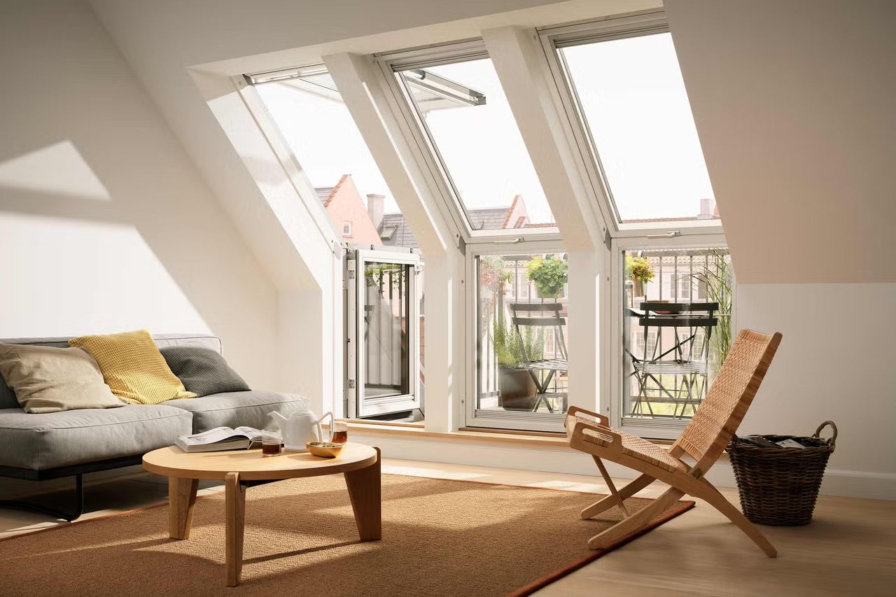 Svetlá podkrovná obývacia izba so strešným balkónom Velux pozostávajúcim z troch okenných jednotiek, pohovkou a dreveným nábytkom.