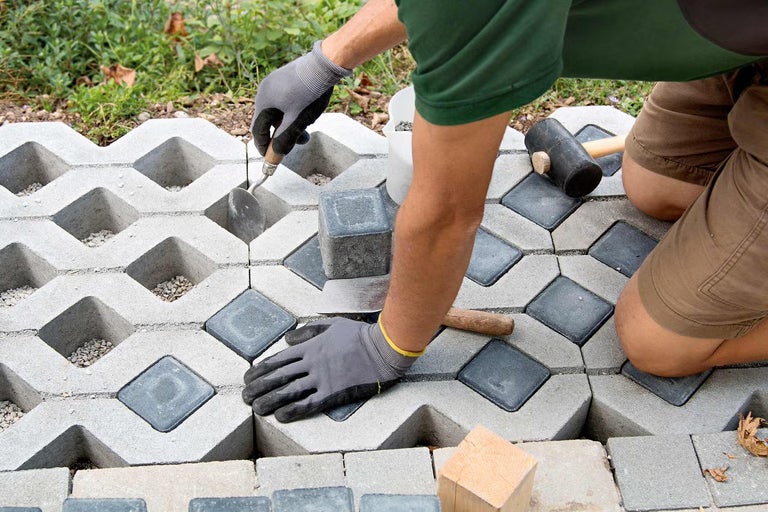 Osoba ukladá malú dlažbu do otvorov betónovej zatrávňovacej dlažby pomocou murárskej lyžice a gumového kladiva.
