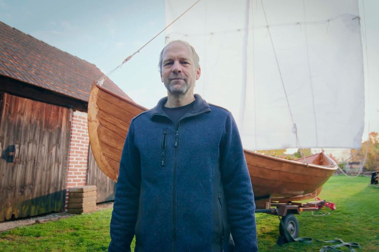 Muž stojí pred drevenou loďou s plachtou na prívese