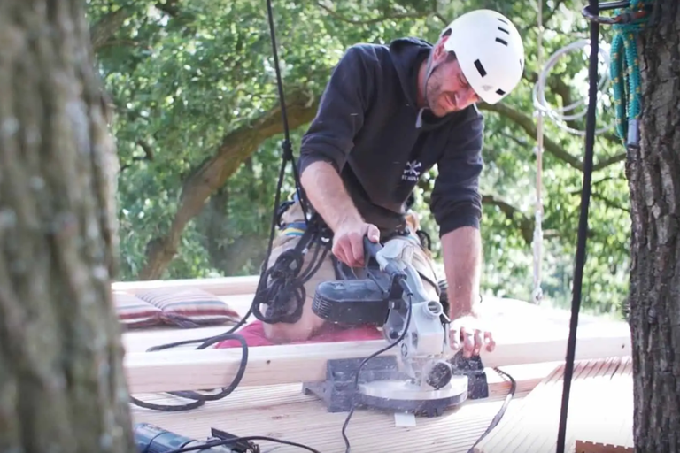 Muž s prilbou reže drevo pokosovou pílou v domčeku na strome