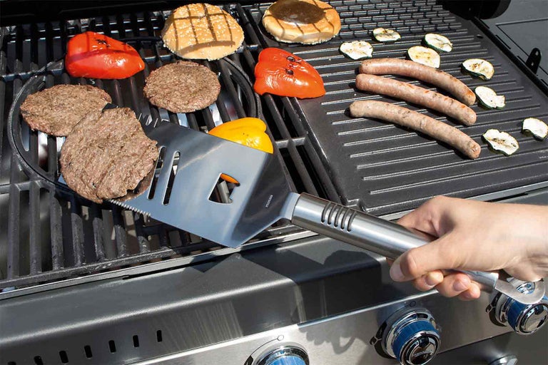 Na grile ležia hamburgery, klobása, paprika a cuketa, zatiaľ čo osoba obracia papriku grilovacou obracačkou.