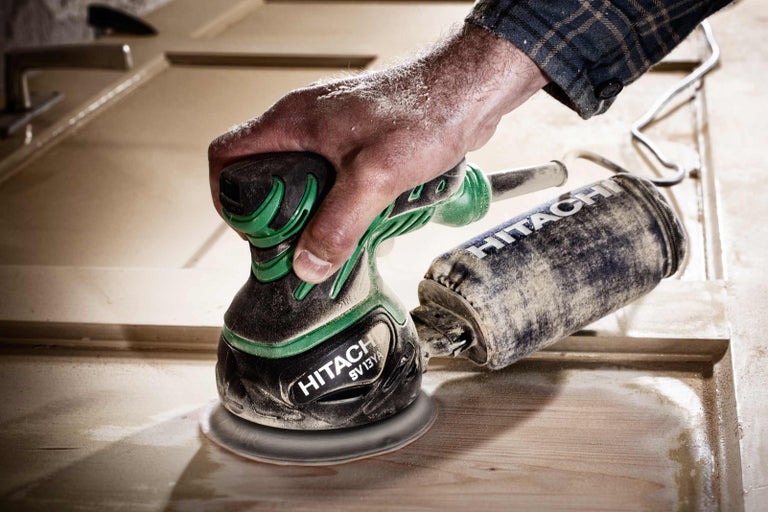 Muž brúsi drevené dvere pomocou excentrickej brúsky značky Hitachi.