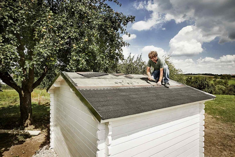 Muž upevňuje bitúmenové šindle na streche záhradného domčeka
