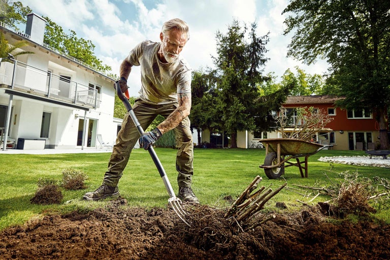 Muž pracuje s rýľovacími vidlami v záhrade, v pozadí je dom a fúrik.