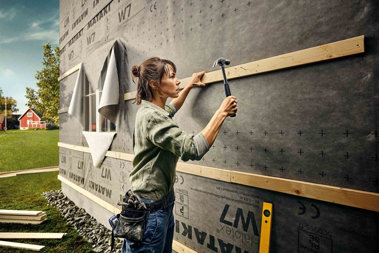 Žena pripevňuje drevené laty kladivom na stenu domu s vetruodolnou fóliou.